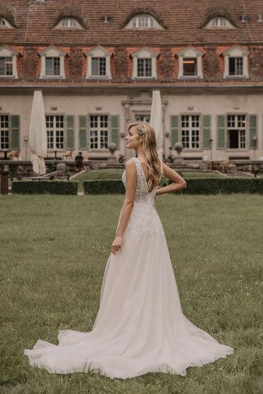 Eine Braut in einem schlichten, aber stilvollen Brautkleid steht auf einer grünen Wiese vor einem alten Herrenhaus mit traditioneller Architektur.