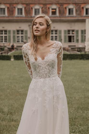 Eine Braut mit einem eleganten, langärmeligen Brautkleid aus Spitze steht vor einem historischen Herrenhaus, schaut nachdenklich zur Seite.